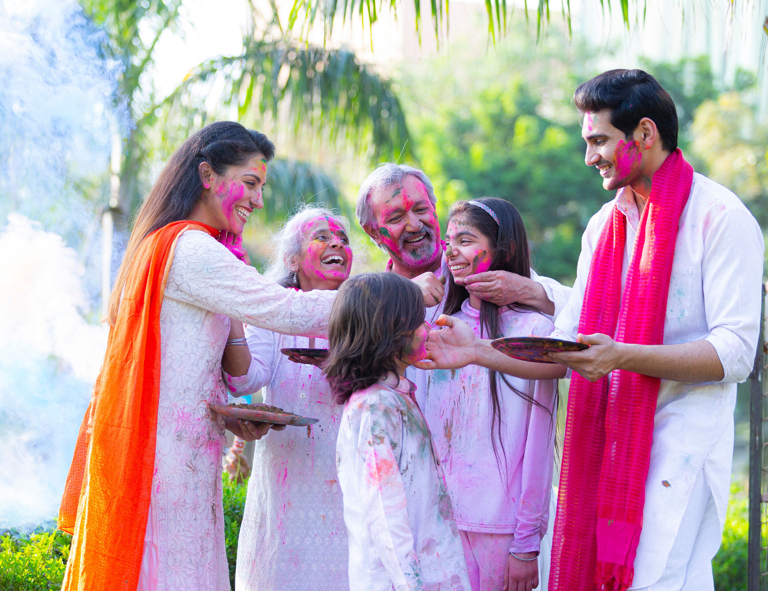 Family celebrating Holi outdoors, smiling as they smear pink gulal on each other, dressed in white ethnic outfits with bright dupattas and color plates.